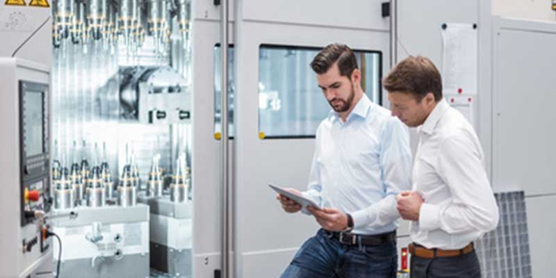 Zwei Männer in weißen Hemden inspizieren eine große Maschine. Einer der Männer hält ein Tablet in der Hand.